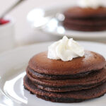 A stack of three chocolate pancakes topped with a swirl of whipped cream sits on a white plate, with another plate of pancakes and a small dish of berry sauce blurred in the background.