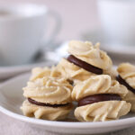 A plate of sandwich cookies with swirled vanilla biscuits and a chocolate filling, with blurred white teacups and more cookies in the background.