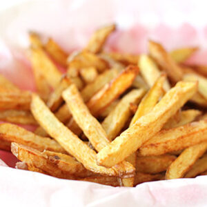 A basket lined with pink and white paper holds a serving of golden, crispy French fries. The fries are cut thin and appear freshly cooked, with a light brown, slightly uneven texture.