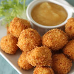 A plate of golden-brown, breaded and fried balls served with a small dish of creamy dipping sauce and a garnish of fresh greens on the side.
