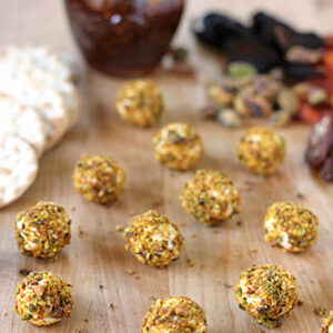 Small balls coated in crushed pistachios are scattered on a wooden surface, with crackers, a jar of fig spread, whole pistachios, and dried fruits blurred in the background.
