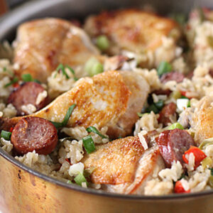 A skillet filled with cooked rice, browned chicken pieces, sliced sausage, chopped green onions, and red bell peppers. The dish looks savory and well-seasoned.