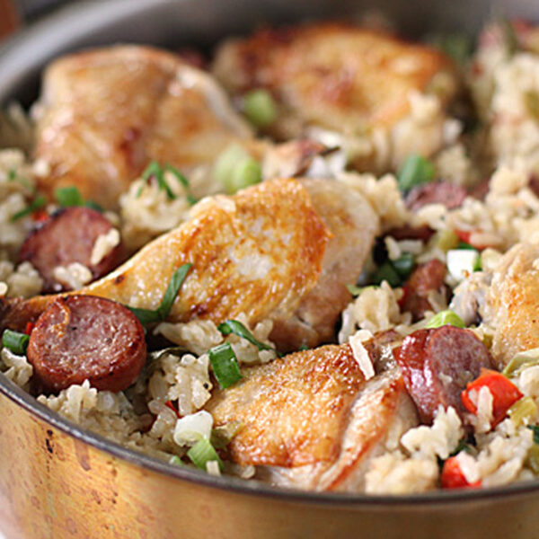 A skillet filled with cooked rice, browned chicken pieces, sliced sausage, chopped green onions, and red bell peppers. The dish looks savory and well-seasoned.