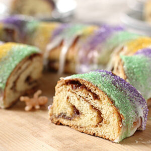 A sliced king cake with colorful sugar in purple, yellow, and green on top, showing a cinnamon swirl inside. A small plastic baby figurine is placed on the wooden surface near the cake.