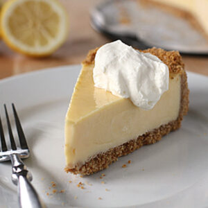 A slice of lemon pie with a graham cracker crust and a dollop of whipped cream sits on a white plate with a fork; lemon halves and the rest of the pie are in the background.