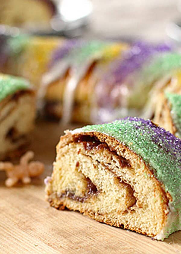 A sliced King Cake with colorful green, purple, and yellow sugar on top and white icing, with a small plastic baby figurine next to a slice on a wooden surface.