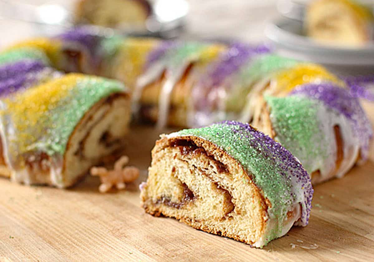 A sliced King Cake with colorful green, purple, and yellow sugar on top and white icing, with a small plastic baby figurine next to a slice on a wooden surface.