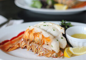 A lobster tail served on a white plate with melted butter in a small cup, garnished with chopped herbs and a lemon wedge, with greens in the background.