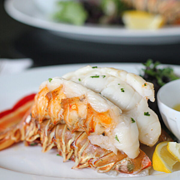 A lobster tail served on a white plate with melted butter in a small cup, garnished with chopped herbs and a lemon wedge, with greens in the background.