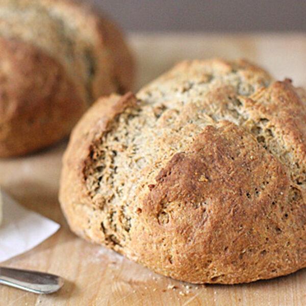 A round loaf of rustic brown bread sits on a wooden surface next to a slab of butter and a butter knife, with another loaf blurred in the background.