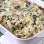 A white baking dish filled with a creamy vegetable casserole, topped with melted cheese and chopped herbs, sits on a green surface with a spoon and a white napkin beside it.