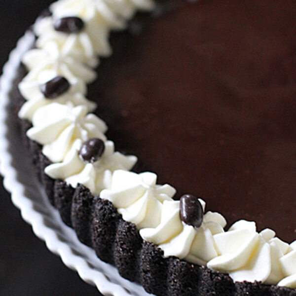 A chocolate tart on a white plate, topped with a ring of piped whipped cream and decorated with chocolate-covered coffee beans around the edge.