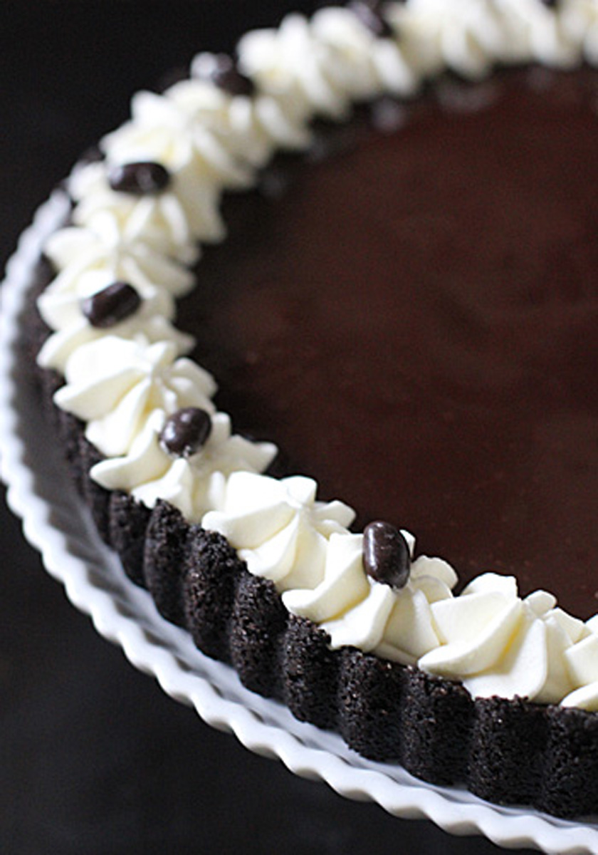A chocolate tart on a white plate, topped with a ring of piped whipped cream and decorated with chocolate-covered coffee beans around the edge.