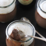 Three small glass jars filled with chocolate pudding topped with whipped cream, one with a spoon inside. A bottle and cap of Guinness beer are in the background.