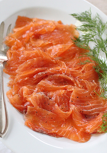 Thin slices of cured salmon arranged on a white plate, garnished with fresh dill and cracked black pepper, with a serving fork on the side.