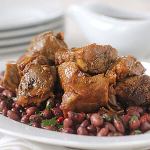 A white plate filled with chunks of cooked, seasoned meat served on a bed of red beans mixed with herbs, with a white gravy boat and stacked plates in the background.