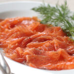 Thinly sliced cured salmon is arranged on a white plate, garnished with fresh dill and cracked pepper. A fork and spoon rest beside the salmon, ready for serving.