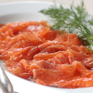 Thinly sliced cured salmon is arranged on a white plate, garnished with fresh dill and cracked pepper. A fork and spoon rest beside the salmon, ready for serving.
