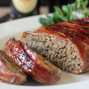 A bacon-wrapped meatloaf sits on a white plate, with two thick slices cut and slightly separated from the loaf. Green herbs garnish the background. The meatloaf has a shiny, glazed surface.