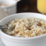 A close-up of a white bowl filled with oatmeal, with a spoon resting inside. The oatmeal appears creamy and slightly textured, and there’s a glass of orange juice and a cup in the blurred background.