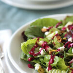A close-up of a fresh green salad with leafy greens, nuts, crumbled cheese, and a vibrant magenta dressing on a white plate, with another plate blurred in the background.