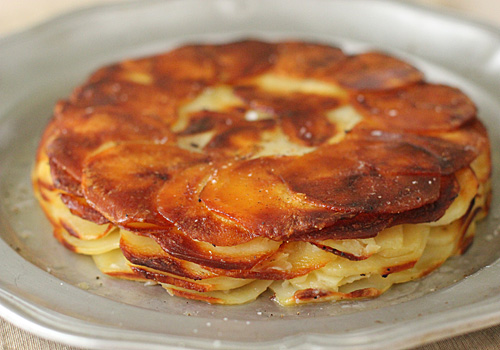 A golden-brown potato dish with thin, overlapping slices baked in a circular pattern on a silver plate, with crispy edges and a slightly glossy surface.