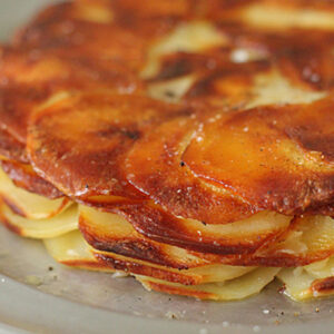 A close-up of a golden-brown potato galette, showing thin, crispy layers of sliced potatoes baked together and neatly arranged in a circular pattern on a metal plate.