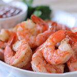A plate of cooked, seasoned shrimp is served with a small bowl of cocktail sauce in the background. The shrimp are pink, plump, and garnished with ground pepper.
