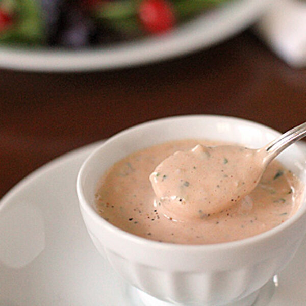 A small white bowl filled with creamy pink salad dressing, garnished with herbs, sits on a white plate. A spoon rests in the bowl, with some dressing on it. A blurry salad appears in the background.