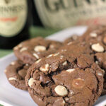 A close-up of chocolate cookies with white and milk chocolate chips on a white plate, with bottles of Guinness beer blurred in the background.