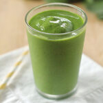 A glass filled with a thick green smoothie sits on a white napkin, with a yellow-striped straw lying beside it on a wooden surface.