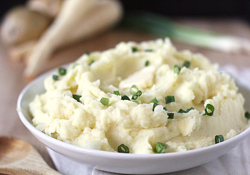 A bowl of creamy mashed potatoes topped with chopped green onions, with a pat of melting butter in the center. Parsnips and a wooden spoon are visible in the blurred background.