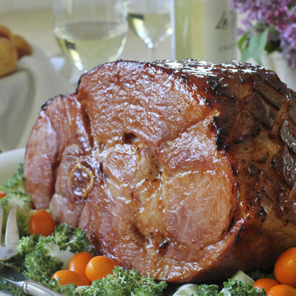 A glazed baked ham sits on a white platter, garnished with leafy greens and orange cherry tomatoes. In the background are dinner rolls, two glasses of white wine, and a vase of purple flowers.