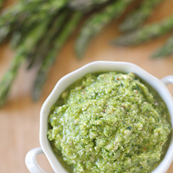 A white bowl filled with green asparagus pesto sits on a wooden surface, with fresh asparagus spears blurred in the background.