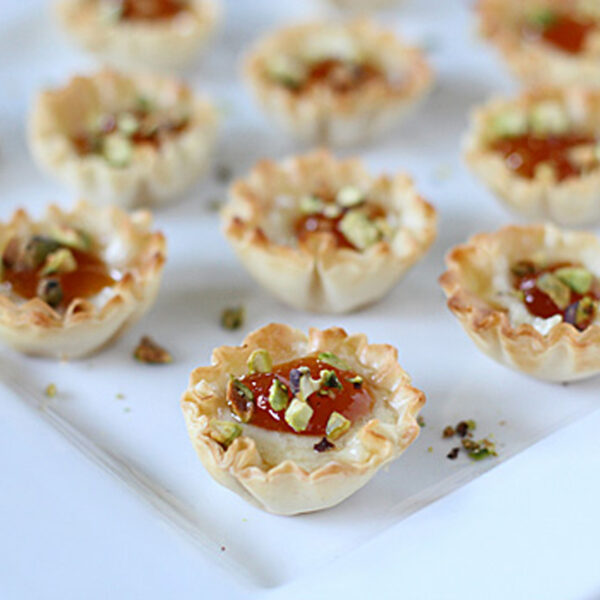 Mini phyllo pastry cups filled with a creamy mixture, topped with a dollop of jam and chopped pistachios, arranged on a white square plate.