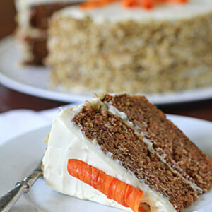 A slice of carrot cake with cream cheese frosting and a decorative carrot made of frosting sits on a plate in front of a whole carrot cake in the background.