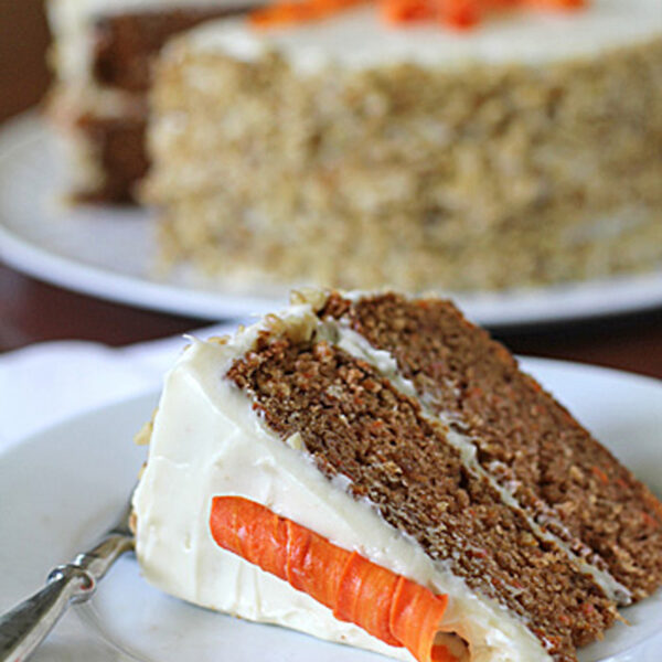 A slice of carrot cake with cream cheese frosting and a decorative carrot made of frosting sits on a plate in front of a whole carrot cake in the background.