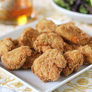 A white plate filled with golden-brown, crispy chicken nuggets sits on a patterned tablecloth, with a salad in a bowl and a drink visible in the background.