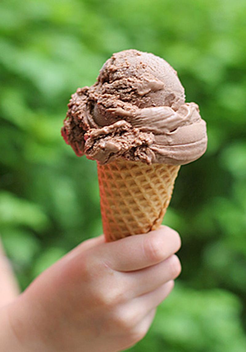 A hand holding a waffle cone with a scoop of chocolate ice cream against a blurred green background.