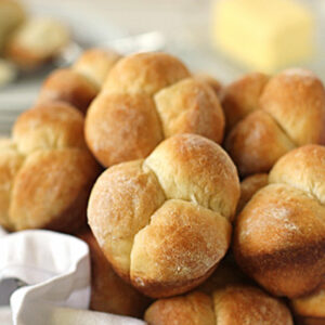 A basket filled with golden brown dinner rolls, with a pat of butter and a plate in the blurred background. The rolls look freshly baked and have a slightly crisp, browned crust.
