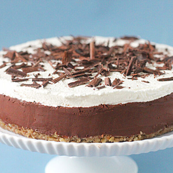 A layered chocolate and cream cake sits on a white cake stand. The cake has a nutty base, a thick chocolate layer, a fluffy white cream topping, and is garnished with chocolate shavings. The background is light blue.