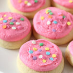 Frosted sugar cookies topped with bright pink icing and colorful round sprinkles, arranged on a white plate.