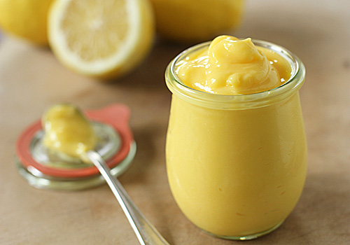 A small glass jar filled with creamy yellow lemon curd, with a spoonful of curd and a halved lemon in the background on a wooden surface.