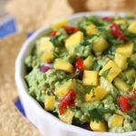 A white bowl filled with guacamole topped with diced mango, red onion, tomato, and cilantro, surrounded by tortilla chips.