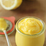 A small glass jar filled with creamy yellow lemon curd sits on a wooden surface, with a spoonful of curd nearby and a halved lemon in the background.