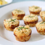 Eight golden-brown, bite-sized appetizers topped with herbs are arranged on a white plate. The food has a crispy texture, and a blurred dish is visible in the background.