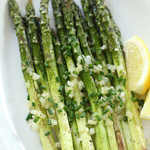 Grilled asparagus spears topped with chopped onions and chives, served on a white plate with lemon wedges on the side.