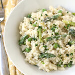 A white bowl filled with creamy risotto, green asparagus pieces, and peas, garnished with chopped herbs. A fork and knife rest on a yellow patterned napkin beside the bowl.