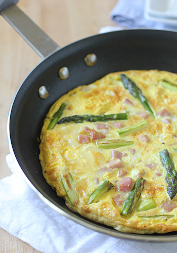 A frittata with pieces of asparagus and ham cooks in a black nonstick skillet, resting on a white cloth on a light wooden surface.