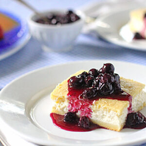 A slice of cheesecake topped with blueberry sauce sits on a white plate, with a fork beside it. In the background, a bowl of blueberry sauce and another dessert plate are visible.
